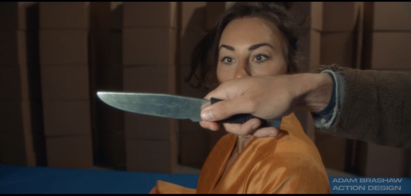 Previs close-up: a knife passes in front of a woman's face in a rehearsal space.